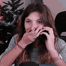 a woman covering her mouth with her hands while talking on a cell phone in front of a christmas tree