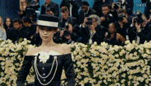 a woman is walking down a runway in front of a crowd of people wearing a hat and pearls .