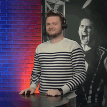 a man wearing headphones and a striped sweater stands in front of a red and blue brick wall