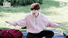 a boy in a pink hoodie sits on a blanket in the grass