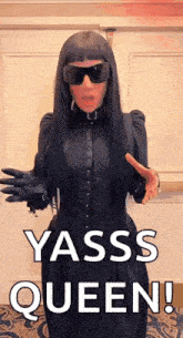 a woman in a black dress and sunglasses is standing in front of a door and says yasss queen !