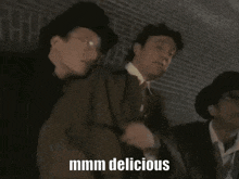 a man in a top hat and glasses is standing in front of a brick wall with the words mmmm delicious above him .