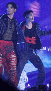 a man in a black jacket and red pants stands next to another man on a stage