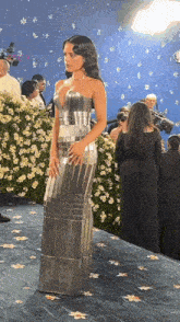 a woman in a silver dress stands in front of a blue wall of flowers