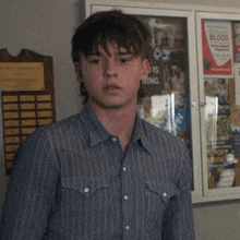 a young man in a striped shirt stands in front of a bulletin board with a poster that says this is hiring