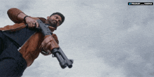 a man in a brown jacket is holding a gun in front of a cloudy sky and the name pranay is on the bottom right