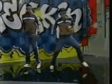 two men are dancing in front of a wall with graffiti on it that says ' rck '