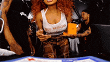 a woman with red hair is wearing a true religion tank top and holding an orange box .