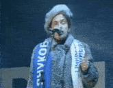 a man singing into a microphone with a scarf around his neck that says ' suvkob '