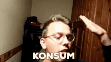 a man wearing glasses salutes in front of a wooden door with the word konsum written on his face .