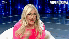 a woman wearing glasses and a pink dress is sitting in front of a sign that says viperissima on it
