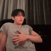 a man in a grey shirt is sitting on a couch with his hand on his chest .