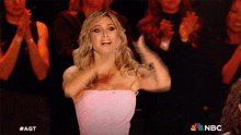 a woman in a pink dress applauds in front of a crowd with the nbc logo in the background