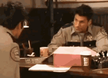 a man in a sheriff 's uniform is sitting at a desk with a cup of coffee