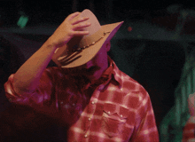 a man wearing a plaid shirt and a cowboy hat covers his face