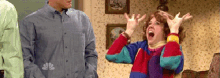 a woman in a colorful sweater is screaming with her hands in the air while a man stands behind her