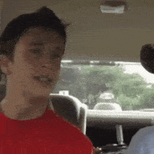 a young man in a red shirt is sitting in the back seat of a car talking to someone .