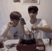two young men are sitting at a table with a pot on a stove while one of them looks at his phone .