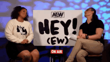 two women sit in front of a sign that says hey