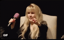 a woman is sitting in a chair holding a microphone and pointing .