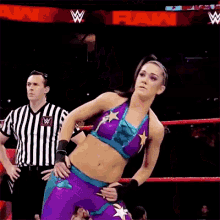 a woman in a purple and blue outfit is standing in a wrestling ring with a referee in the background .