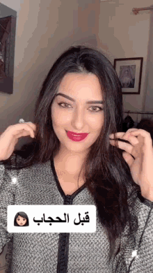 a woman wearing a gray sweater and red lipstick holds her hair in front of a sign that says قبل الحجاب
