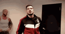 a man in a red and black jacket is standing in a room next to another man .
