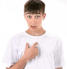 a young man wearing a white t-shirt is pointing at his chest .