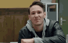 a man in a leather jacket and hoodie is sitting at a table with his arms crossed .