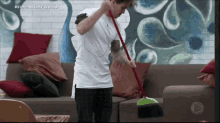 a man in a white shirt is holding a broom in front of a couch with red pillows