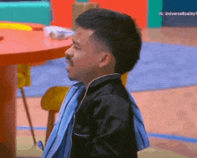 a man wearing a black jacket and a blue towel around his neck is sitting at a table .