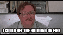 a man with glasses and a mustache is sitting at a desk in front of a shelf of papers .