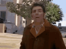 a man in a brown coat is standing in front of a building with a tree in the background .