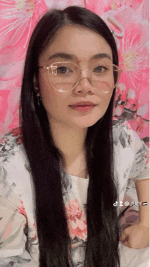 a woman wearing glasses is standing in front of a pink wall with flowers