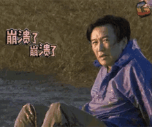 a man in a blue jacket is sitting on the ground laughing with chinese writing behind him .