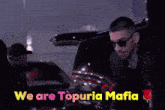 a man in a suit stands in front of a sign that says we are topuria mafia