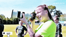 a woman in a green shirt is taking a picture with a wolfe tv logo on the bottom right