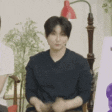 a man in a black shirt is sitting in a chair in front of a lamp and a plant .