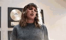 taylor swift is wearing a gray sweater and standing in front of a wall with plaques on it .
