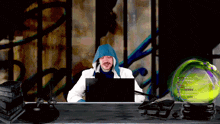 a man in a blue hoodie sits at a desk with a laptop in front of him