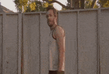 a man in a tank top stands in front of a fence