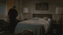 a woman stands next to a man laying on a bed that has a cbs logo on it