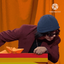 a man wearing a beanie and sunglasses is playing with a paper boat in front of a curtain with the letters k on it