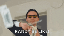 a man in a suit and sunglasses is holding a stack of money and saying `` randy be like ! ''