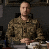 a man in a military uniform is sitting at a desk with his hands folded in front of him