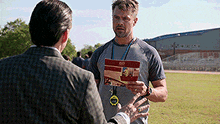a man in a suit is talking to another man in a field while holding a booklet .