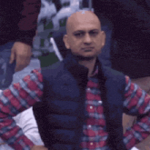 a bald man wearing a plaid shirt and a blue vest looks at the camera