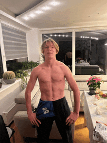 a shirtless man stands in a living room with a blue bag that says ' 85 ' on it