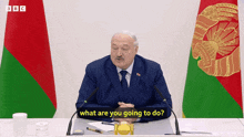 a man in a suit and tie is sitting at a table with microphones and the words what are you going to do behind him