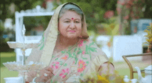 an elderly woman in a floral sari is sitting at a table with a glass of tea .